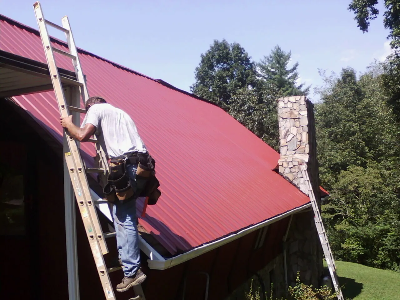 Roofer on a ladder beside a red metal roof and stone chimney — Soffit & Fascia Repair by Roofing Brothers in Canute